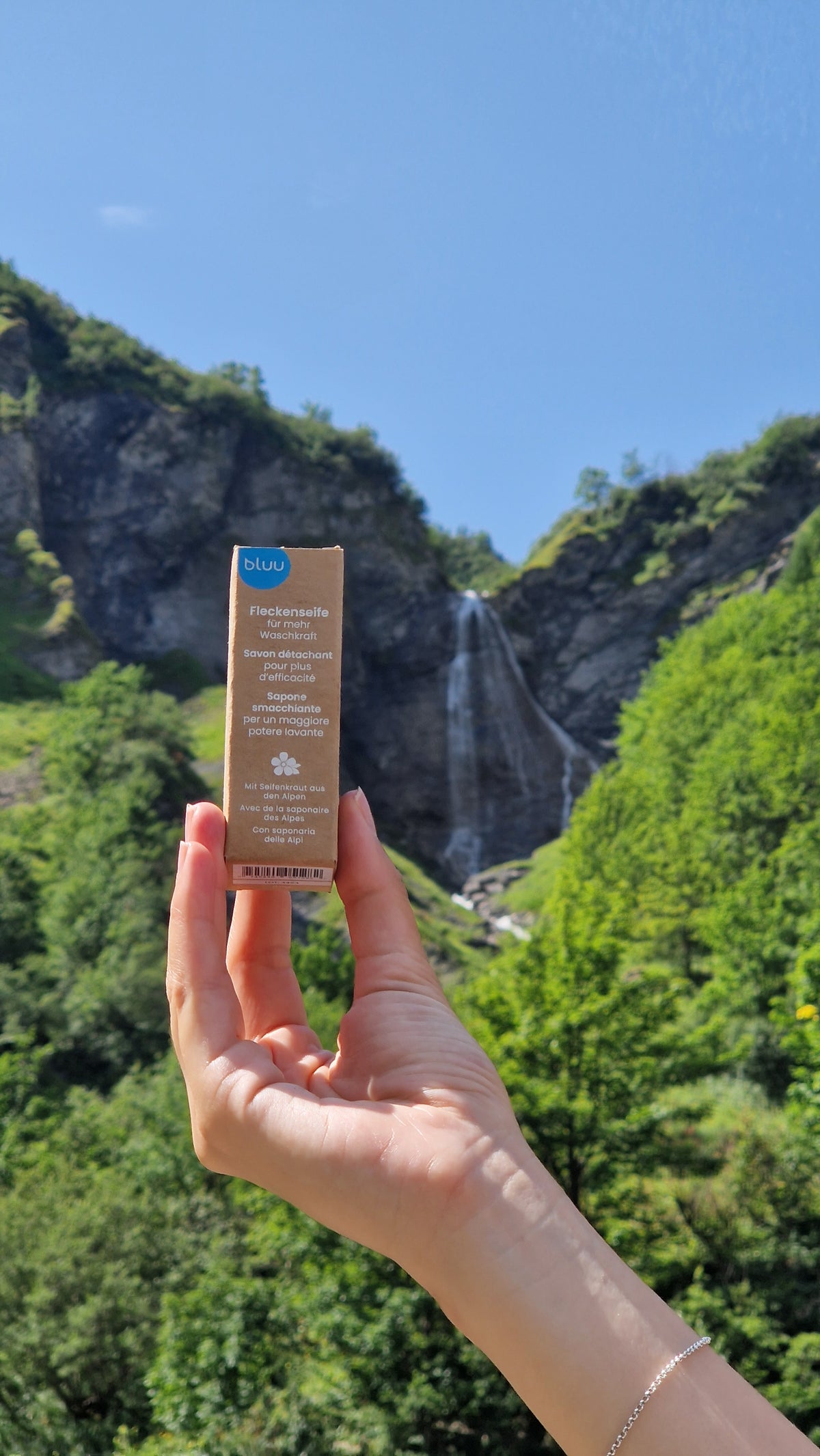 Hand hält eine bluu Waschstreifen-Packung vor einem Wasserfall im Wald