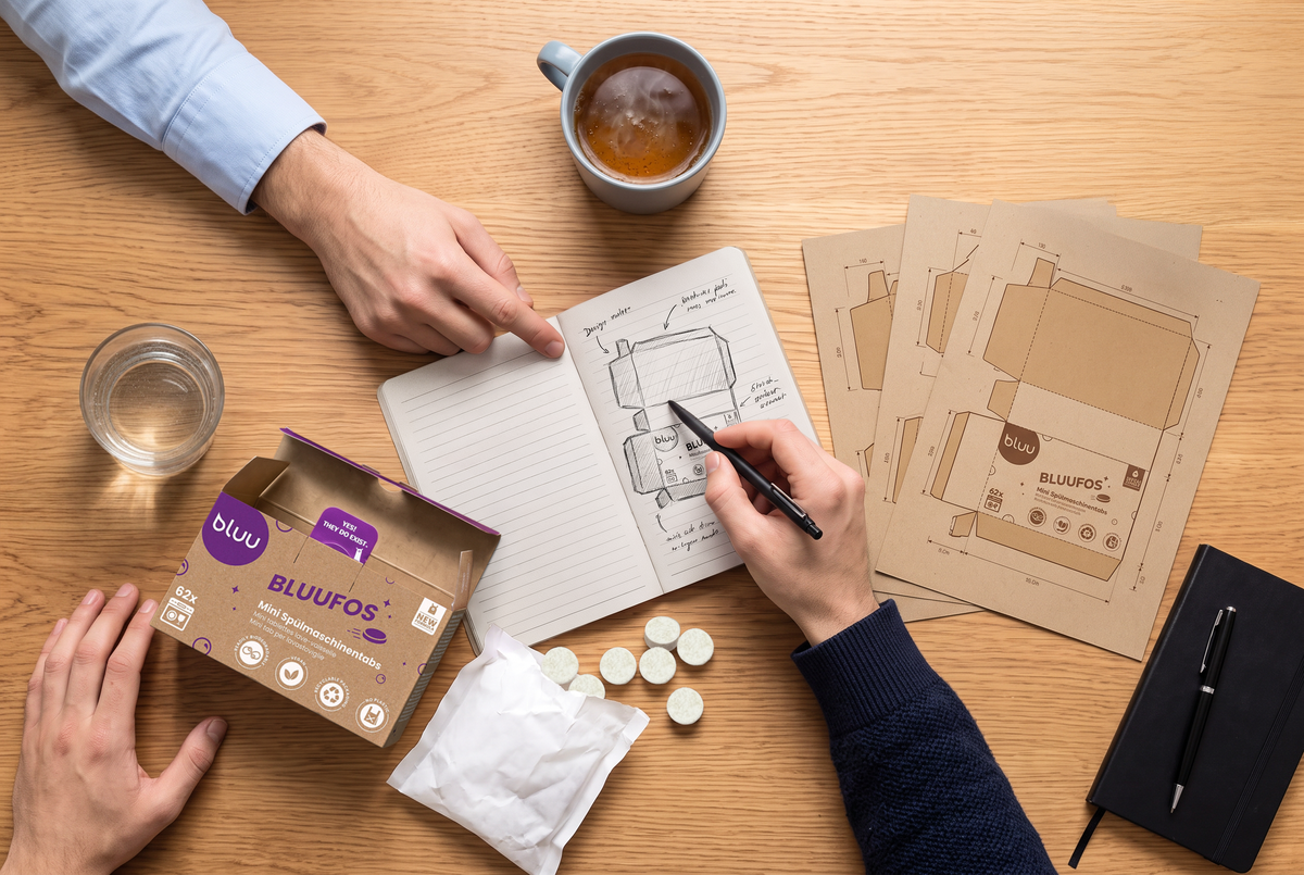 Due mani disegnano una confezione su un quaderno, accanto a una confezione bluufos aperta e fogli di punzonatura per imballaggi