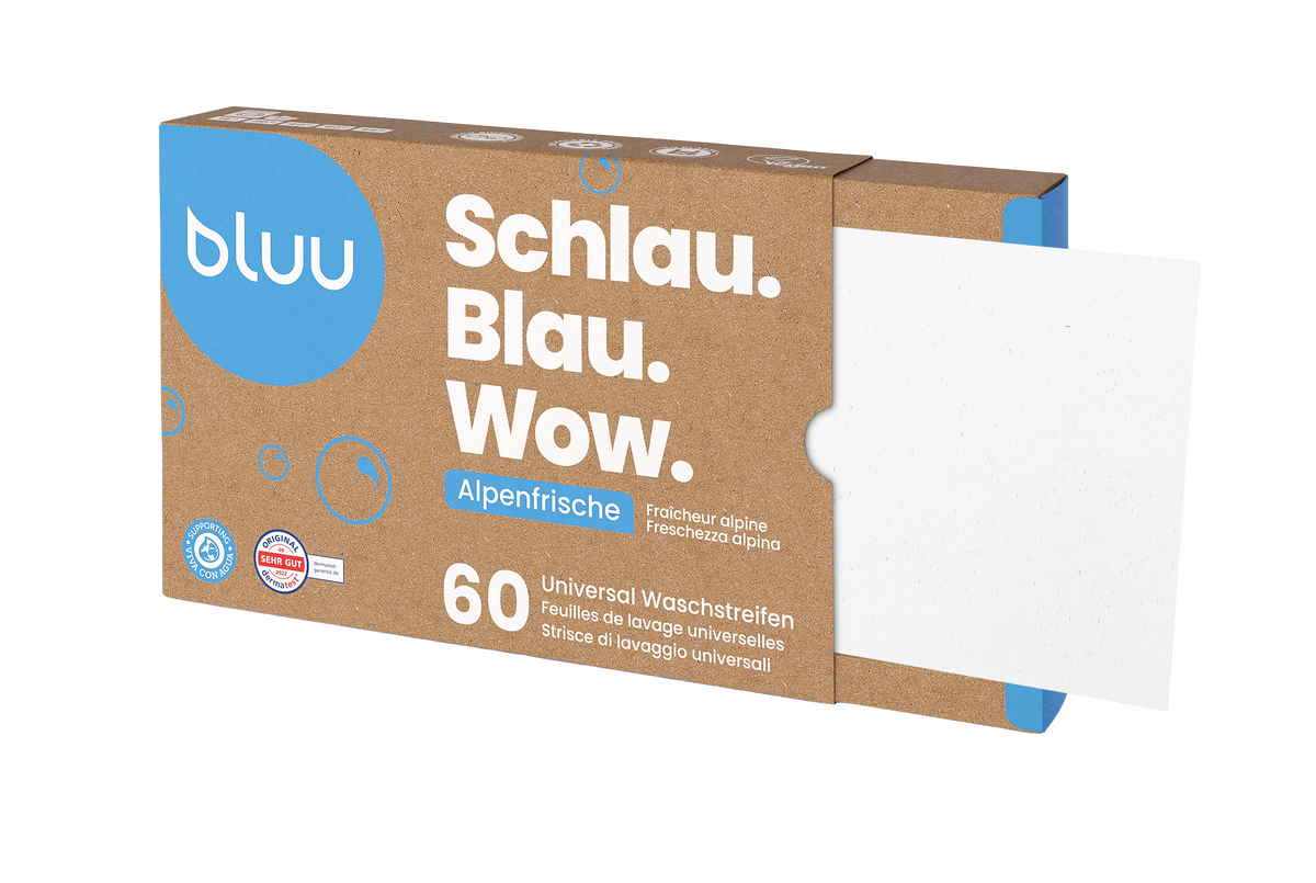 Mutter und Tochter spielen lachend mit einem Wäschekorb voller Handtücher in der Küche, im Hintergrund eine Waschmaschine und eine bluu-Verpackung.
