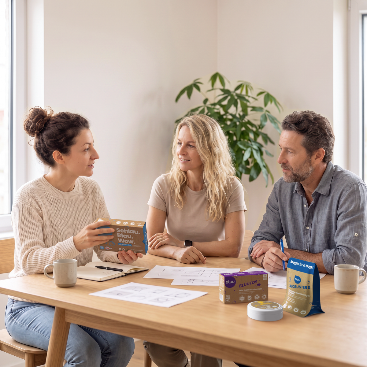 Trois personnes sont assises en conversation autour d'une table, une femme tient un paquet bluu «Schlau. Blau. Wow.», et des bluufos et des calcbusters sont posés sur la table.