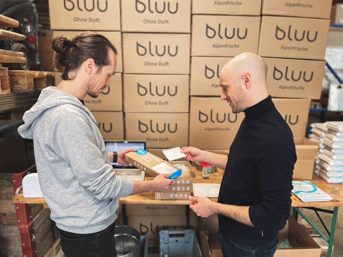 Zwei Männer verpacken bluu-Produkte im Lager vor gestapelten bluu-Kartons